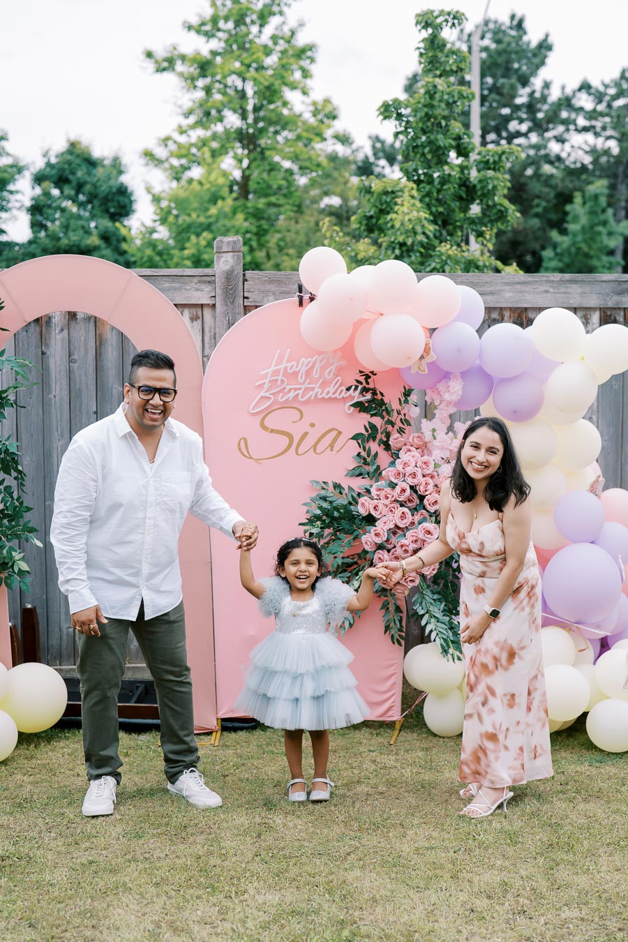 A joyful family celebrating a child's birthday outdoors with pink and purple balloons, a floral backdrop, and a Happy Birthday Sia sign.