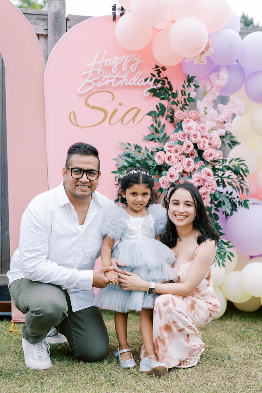 A family celebrating a child's birthday outdoors, with a pink backdrop decorated with flowers and balloons, displaying Happy Birthday Sia. The parents and their daughter are smiling and dressed in festive attire.