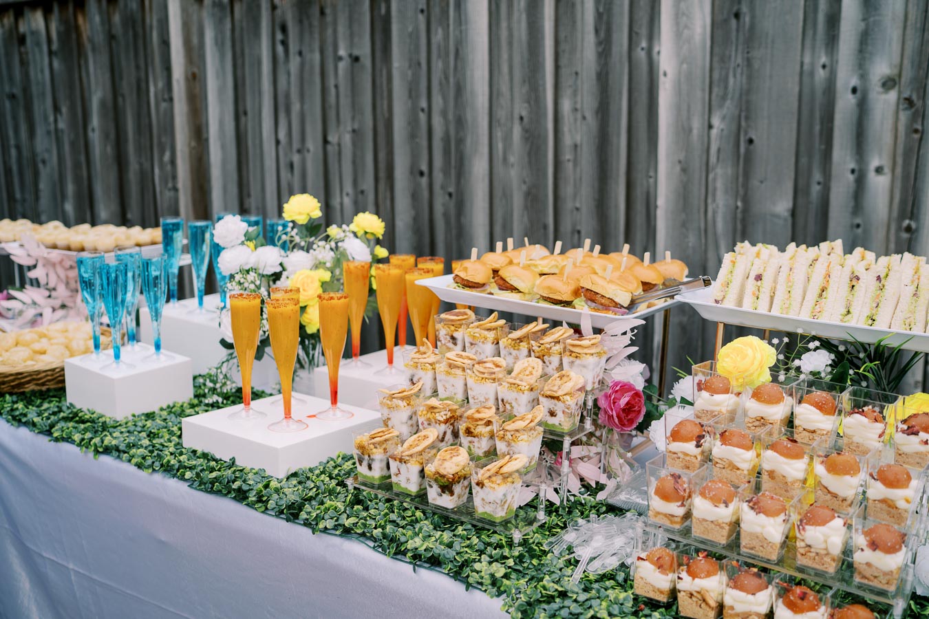 Elegant outdoor buffet table with assorted finger foods, sliders, colorful drinks, and floral decorations, set against a wooden fence background.