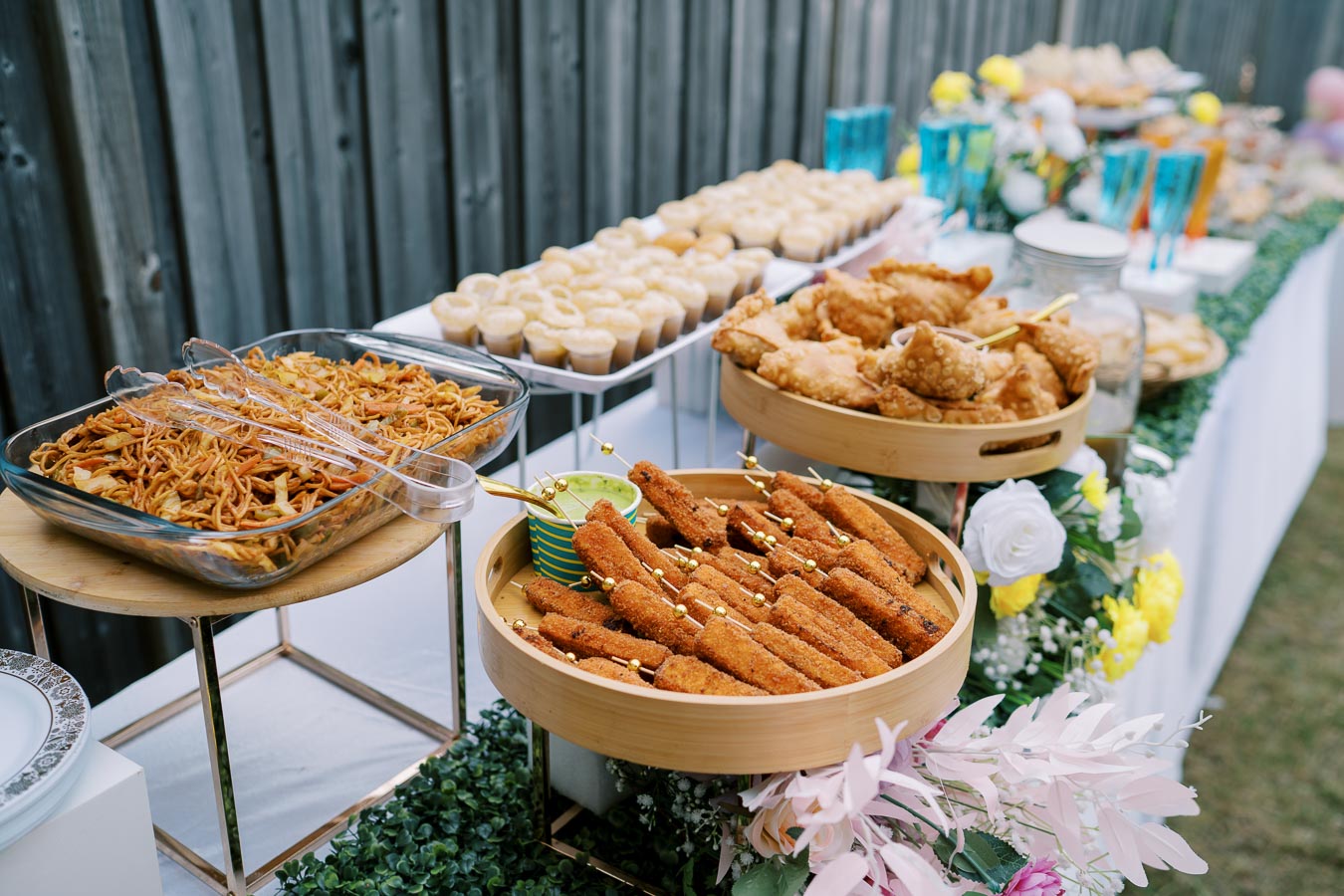 Outdoor buffet table with a variety of appetizers including noodles, fried skewers, and pastries, decorated with flowers, perfect for a garden party or event catering.
