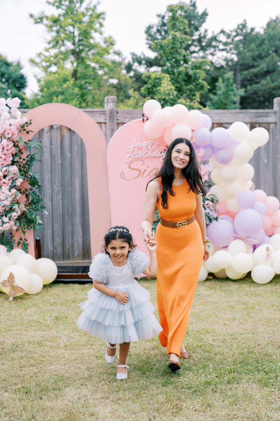 A woman in an orange dress and a young girl in a blue dress holding hands and smiling at an outdoor birthday party with colorful balloon decorations.