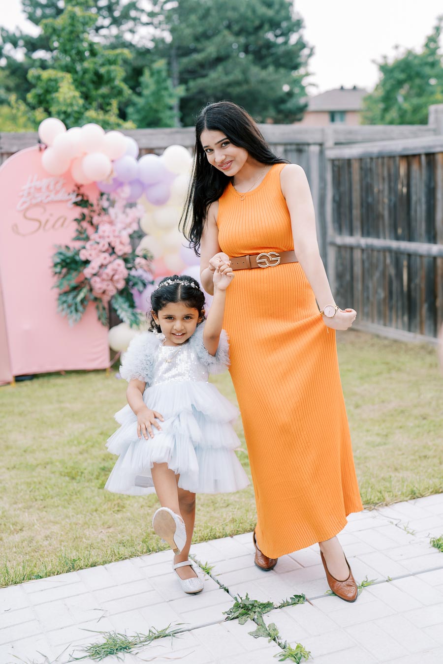 A cheerful young woman in a bright orange dress holds hands with a smiling little girl in a blue frilly dress at an outdoor birthday party, surrounded by colorful balloons and pink flower decorations.