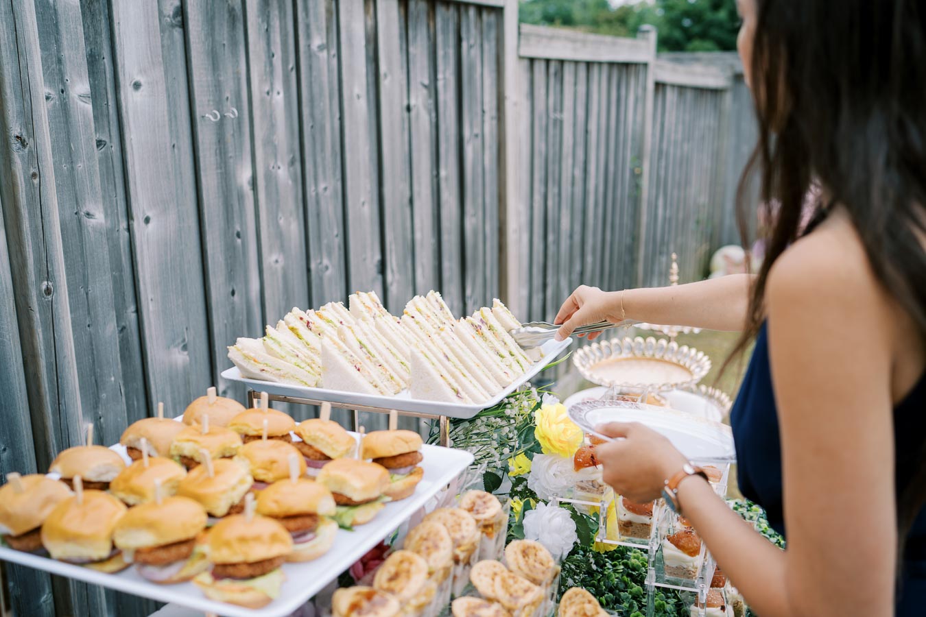 A person serving themselves at an outdoor buffet featuring a variety of sandwiches, sliders, and hors d'oeuvres on a wooden table setup against a wooden fence background.