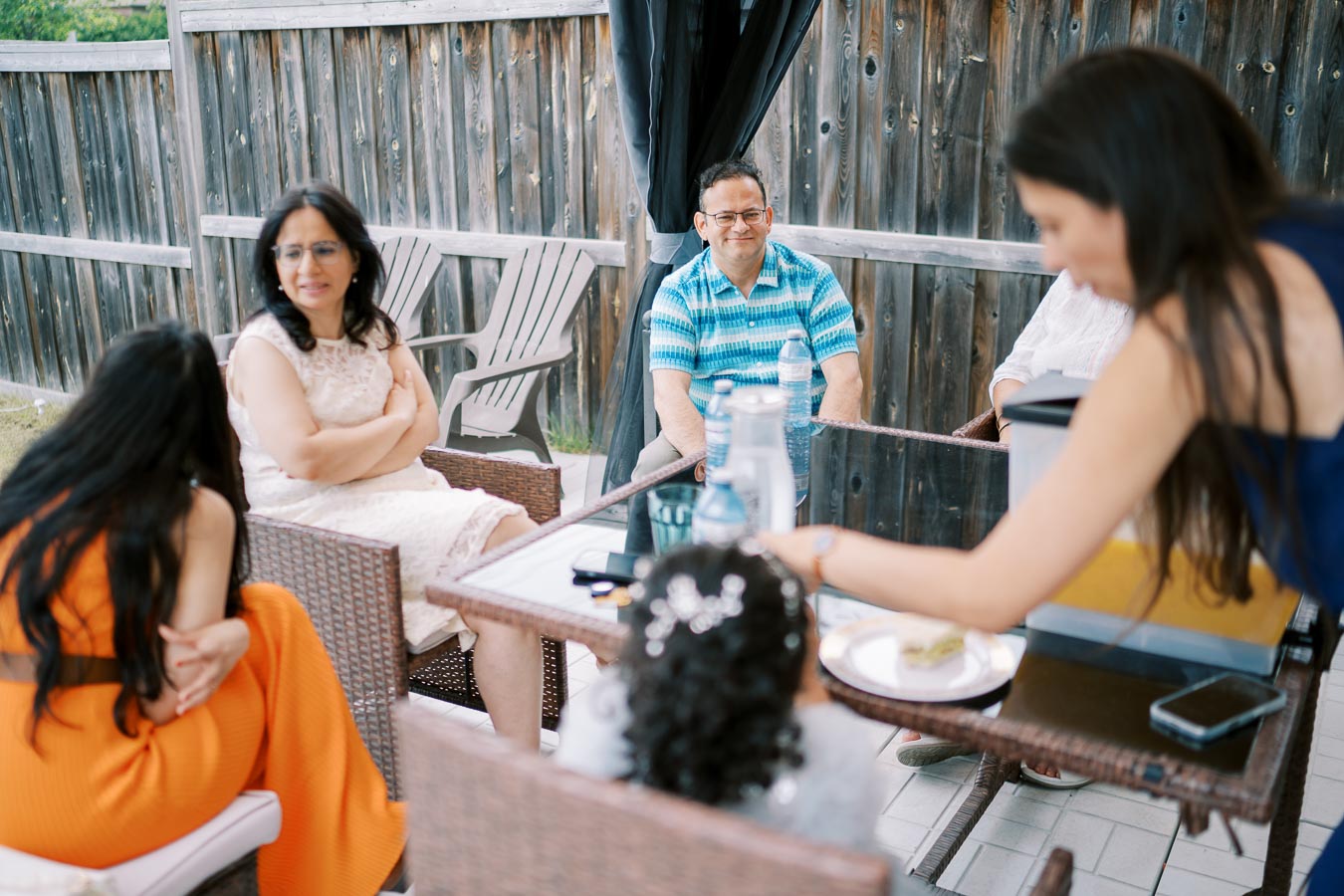 A group of people sitting outdoors around a patio table, engaging in a casual gathering with drinks and snacks, surrounded by a wooden fence.
