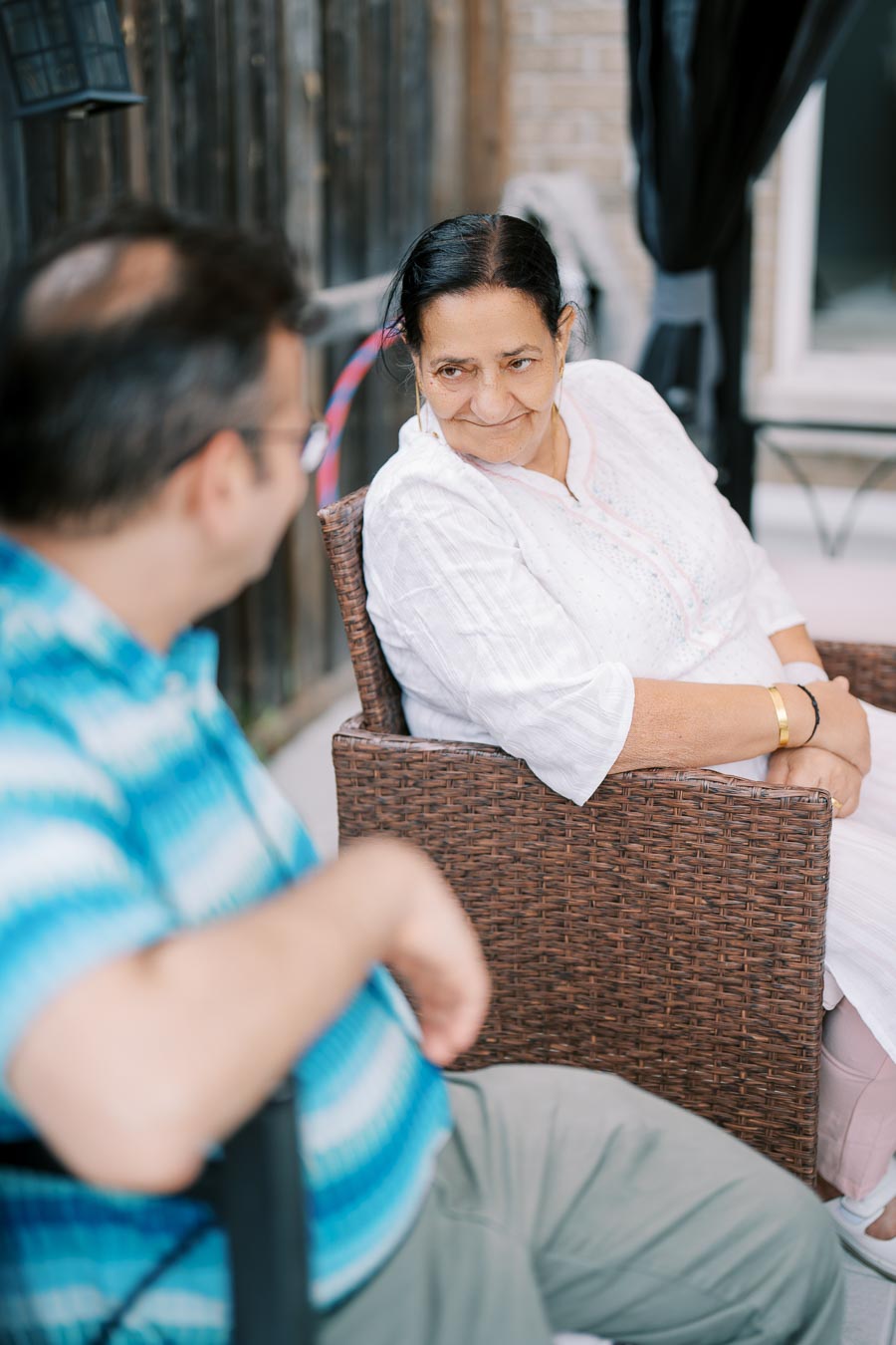 Elderly woman in a white blouse sitting outdoors on a wicker chair, engaging in a conversation with a man in a blue striped shirt.