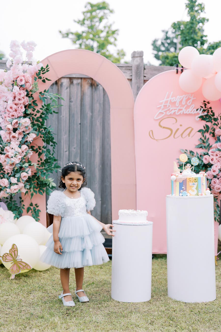 A young girl wearing a blue dress stands smiling at a birthday party, surrounded by pink floral decorations and balloons, with a pink and white cake displayed on a pedestal.
