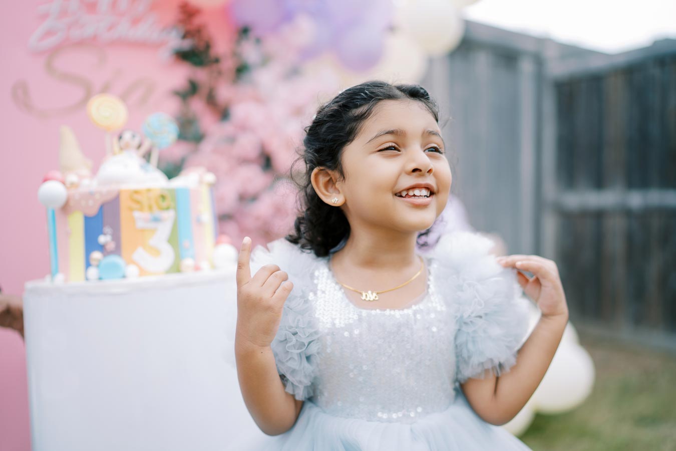 A joyful young girl in a sparkly dress celebrates her third birthday with a colorful cake in the background at an outdoor party.