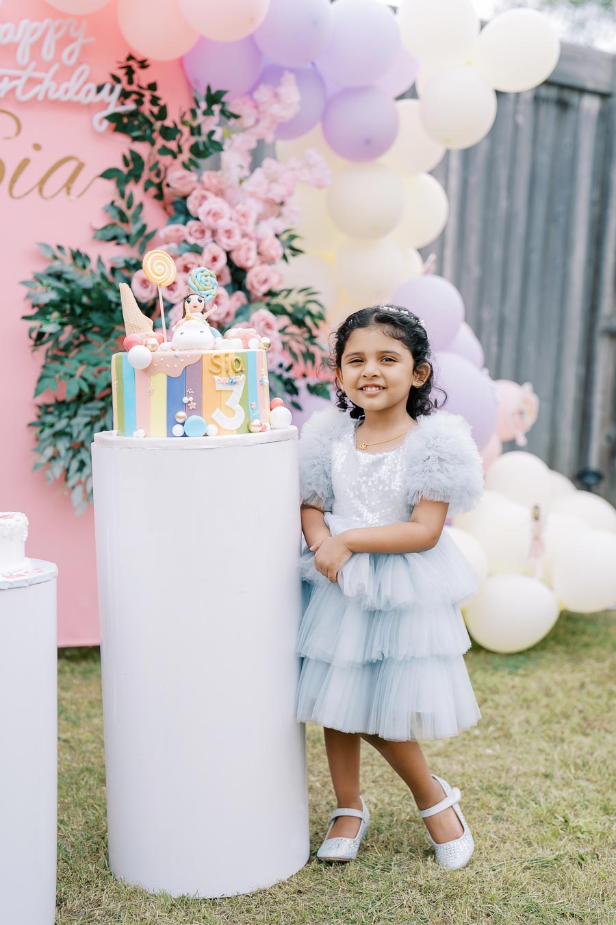 A young girl in a blue dress smiles at her birthday party, standing next to a colorful cake decorated with candy-themed elements. The background features pastel balloons and flowers, creating a festive and cheerful atmosphere.