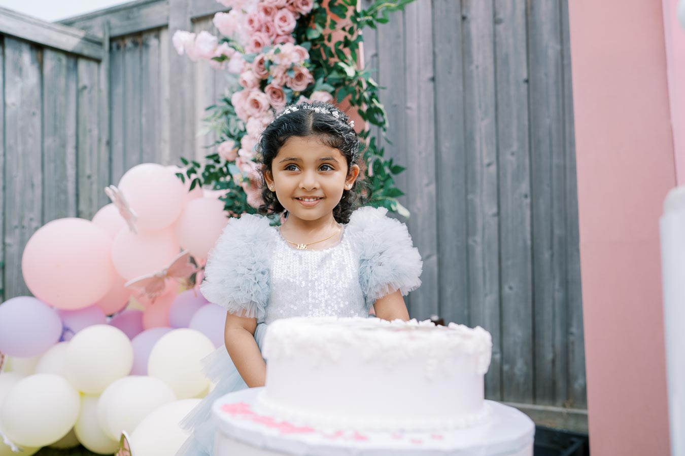 A young girl in a sparkly dress celebrates her birthday outdoors, standing in front of a white frosted cake. She is surrounded by pastel-colored balloons and pink floral decorations.