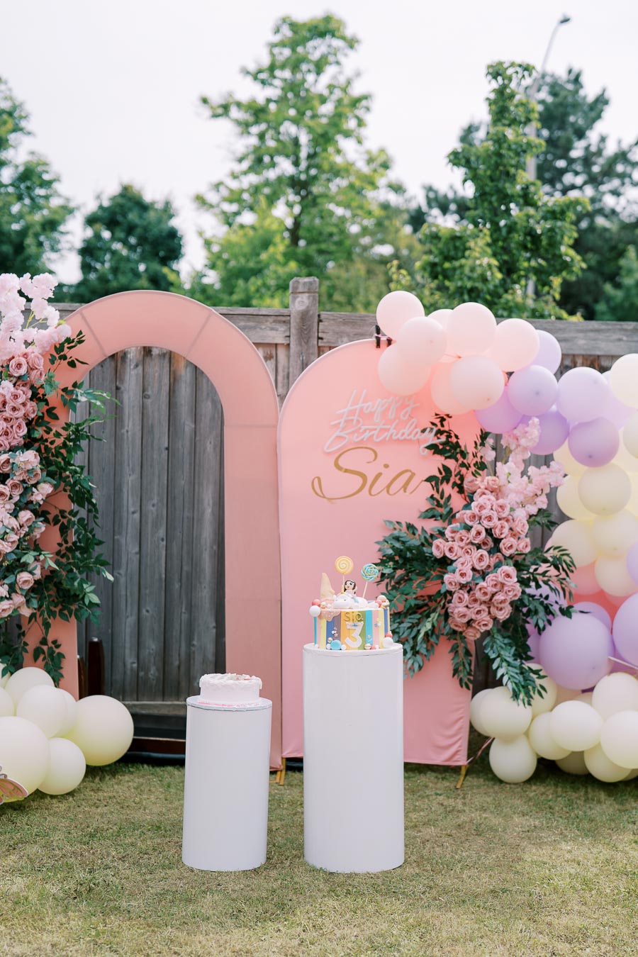 Celebratory backyard birthday setup with two cakes on white pedestals, surrounded by pink and white balloons and floral arrangements, pink arches, and a wooden fence, creating a festive atmosphere for a child’s party.