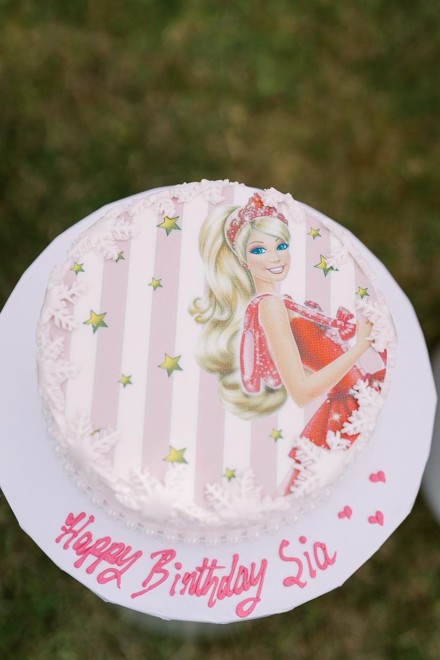 A birthday cake with a pink and white striped design, featuring an illustration of a blonde character in a red dress, adorned with stars and snowflake decorations. The cake is personalized with red icing that reads Happy Birthday Lia on the base. The cake is displayed outdoors on a grassy background.