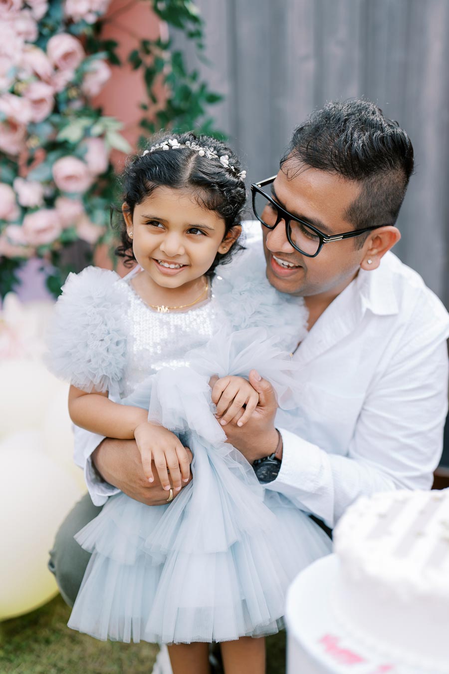 Father and daughter smiling together at a celebration, with the little girl wearing a light blue dress and floral headband, surrounded by a backdrop of pink roses and greenery.