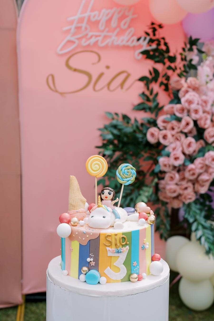 Colorful unicorn-themed birthday cake with lollipop decorations and a figurine topper, set against a pink floral backdrop with a Happy Birthday sign. Perfect for a child's celebration.