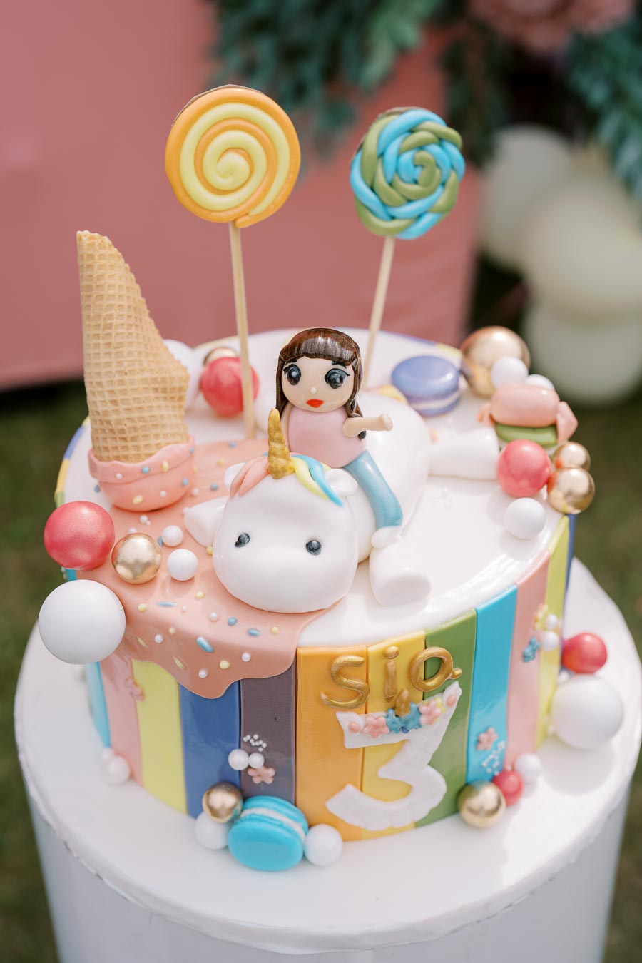 Colorful unicorn-themed birthday cake with a fondant child figure, topped with lollipops, ice cream cone, and decorative candies, celebrating a third birthday.