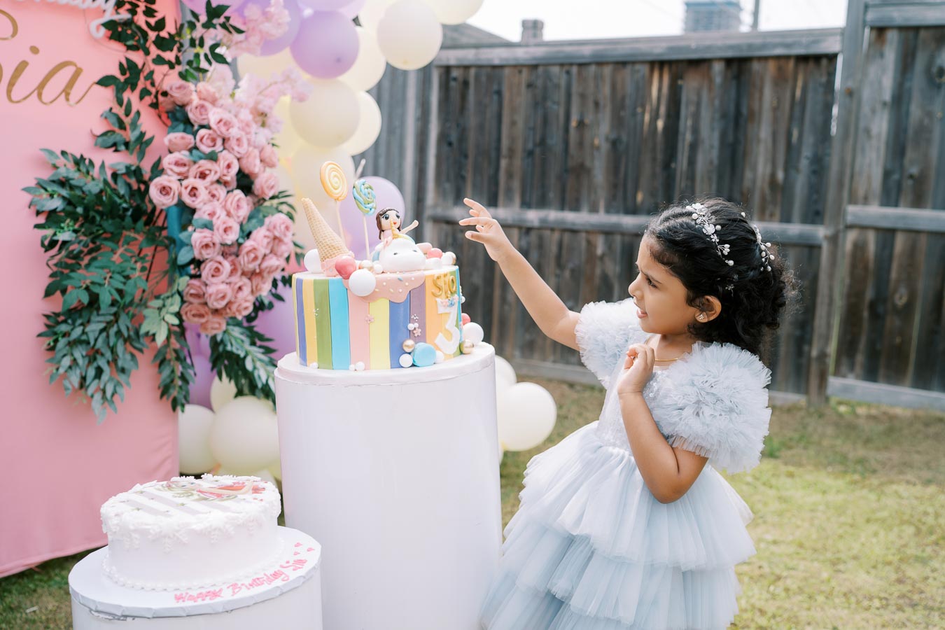 A young girl in a blue dress celebrates at a colorful birthday party outdoors, reaching towards a vibrant birthday cake decorated with pastel stripes and playful toppers, surrounded by pink flowers and balloons.