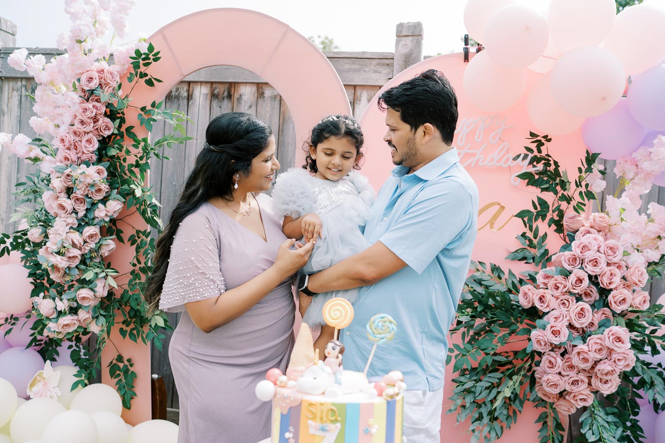 A family celebrates a child's birthday in a beautifully decorated outdoor setting with pink and white balloons, floral arrangements, and a colorful birthday cake. The mother and father smile at their daughter, who wears a fluffy white dress.