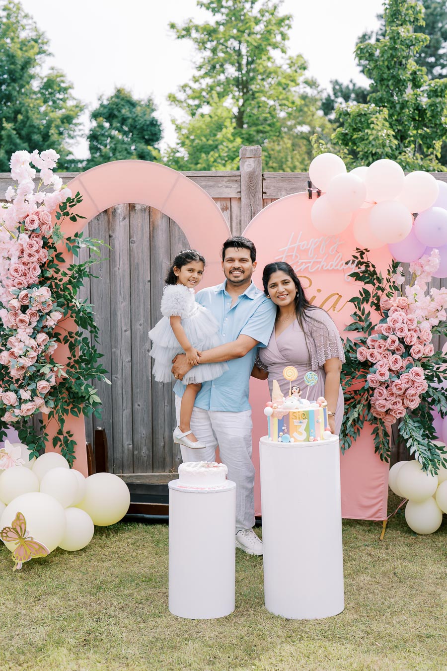 A family celebrating a child's birthday outdoors with pastel-themed decorations, including pink and purple flowers, balloon arrangements, and a colorful birthday cake.