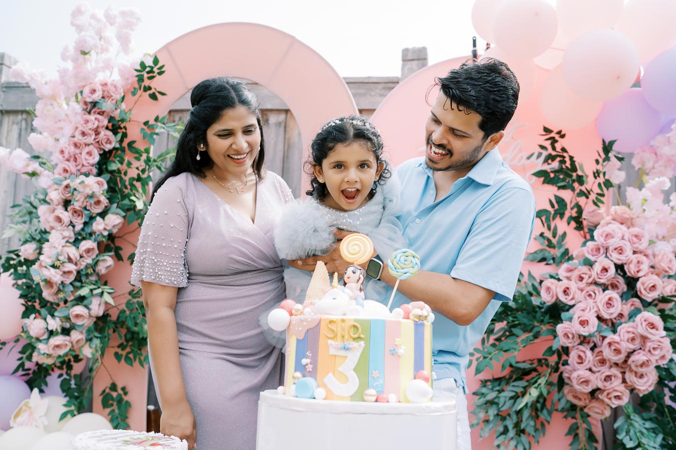 A joyful family celebrating a child's third birthday with a colorful cake and festive balloon decorations, surrounded by pink flowers. The little girl is excitedly held by her parents in a garden setting.
