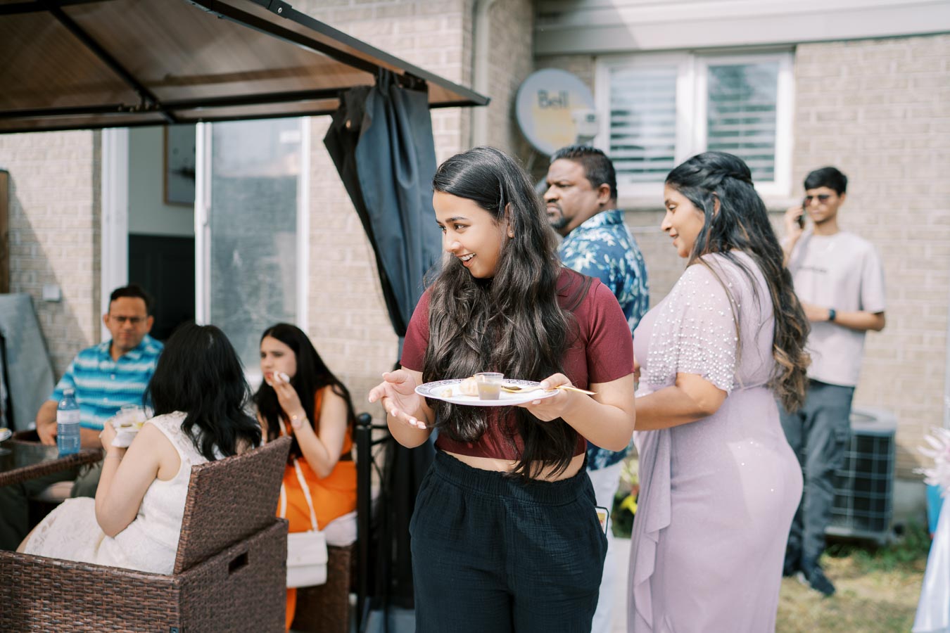 A group of people enjoying an outdoor gathering, with individuals sitting at a table and a woman holding a plate of food, set against a background of a brick house and patio.