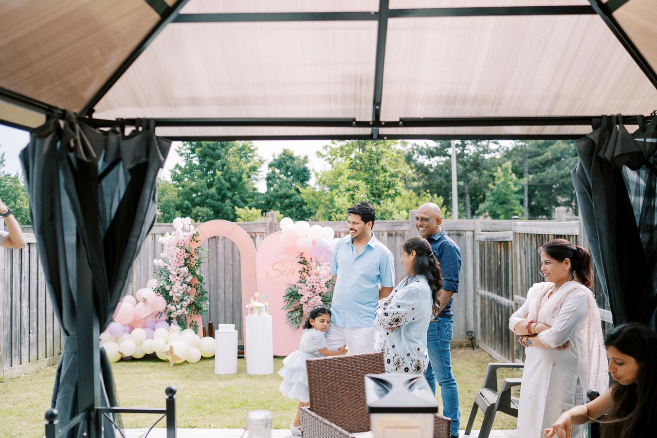 A family gathering in a backyard with decorations, balloons, and a floral backdrop, capturing a joyful moment during a celebration.