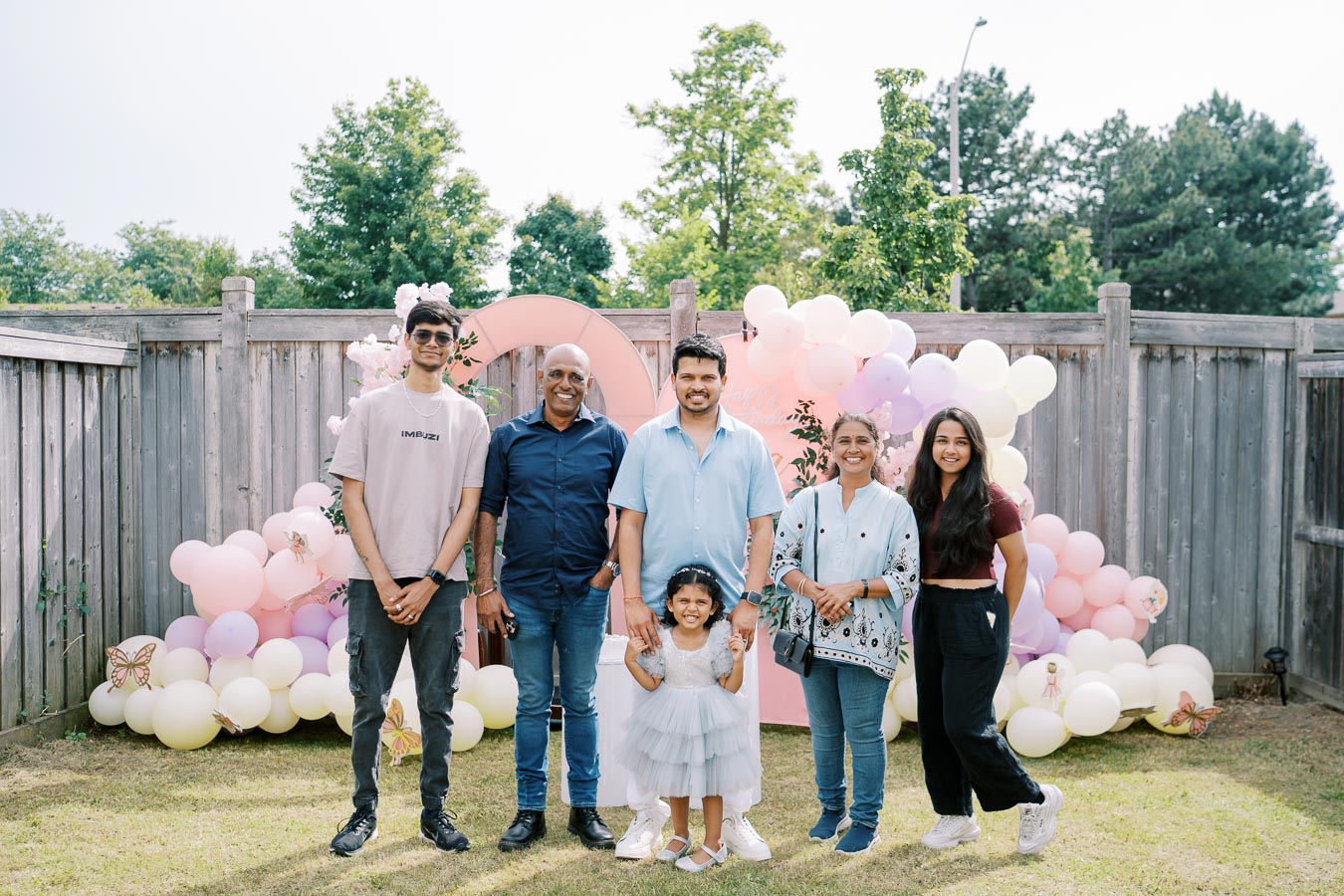 A group of six people smiling outdoors, standing in front of a festive backdrop of pastel balloons and a wooden fence, on a sunny day.