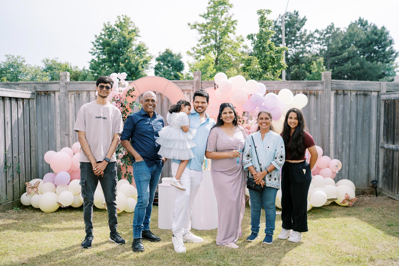 A group of seven people smiling and posing at an outdoor party decorated with pink and white balloons and floral arrangements. Some individuals are dressed casually, while others wear more formal attire. Bright and sunny day, with trees and wooden fence in the background.