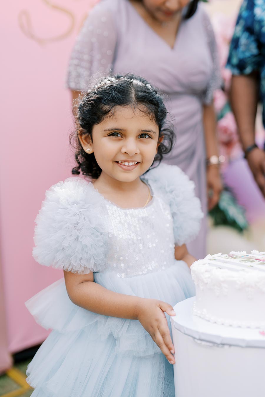 A young girl in a sparkly blue dress stands happily by a white cake, surrounded by family, at a celebration event with pastel décor.