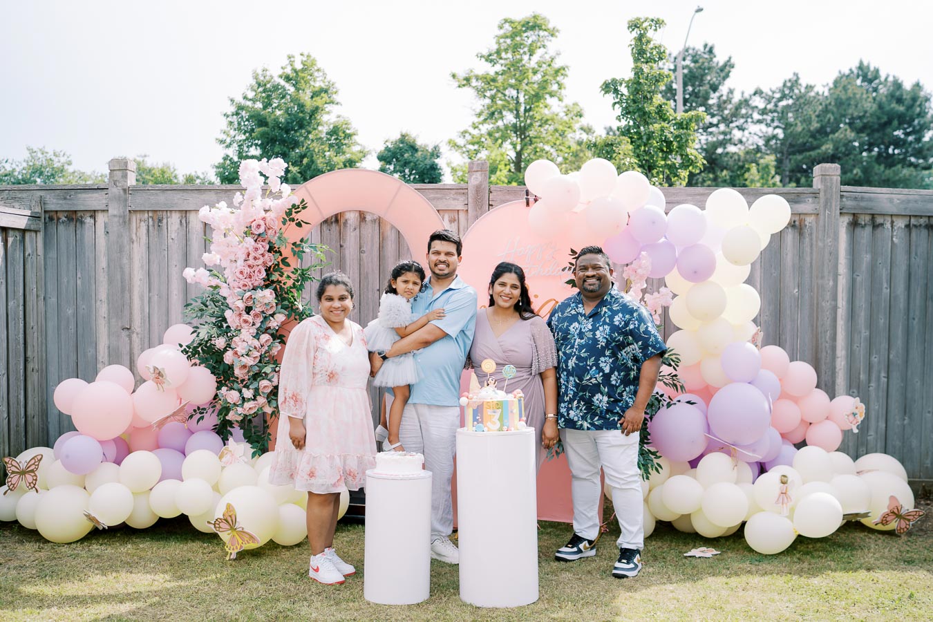 Family celebrating a child's third birthday in a backyard decorated with colorful balloons and floral arrangements, featuring a themed birthday cake and festive atmosphere.