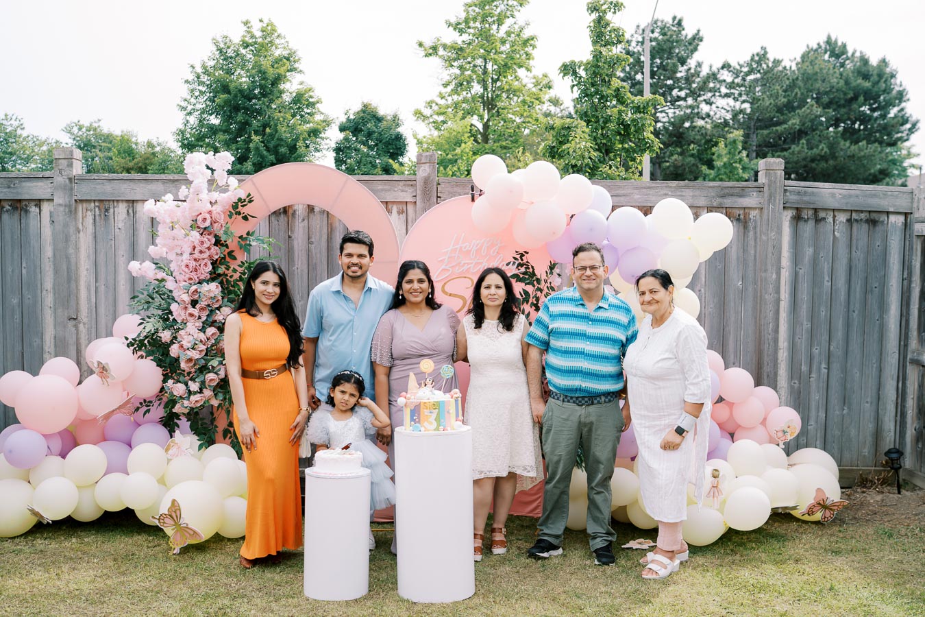 Family gathered outdoors for a 3rd birthday celebration with colorful balloons, a decorative flower arrangement, and a birthday cake, creating a festive atmosphere.