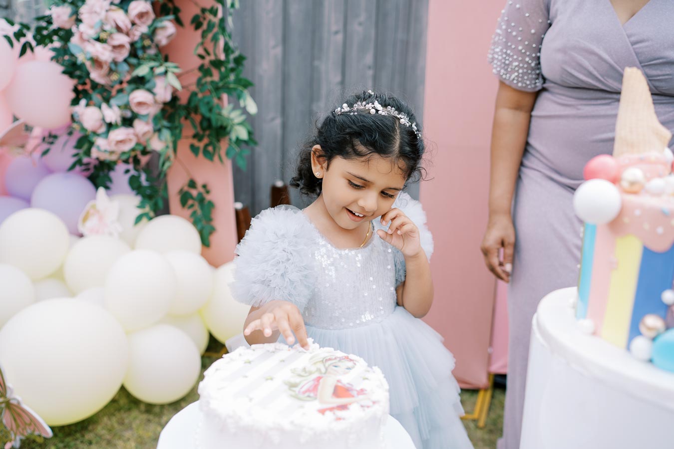 A young girl in a sparkling blue dress, adorned with a floral headband, joyfully touches a decorated cake at a festive outdoor birthday party. Balloons and floral decorations in the background enhance the celebratory atmosphere.