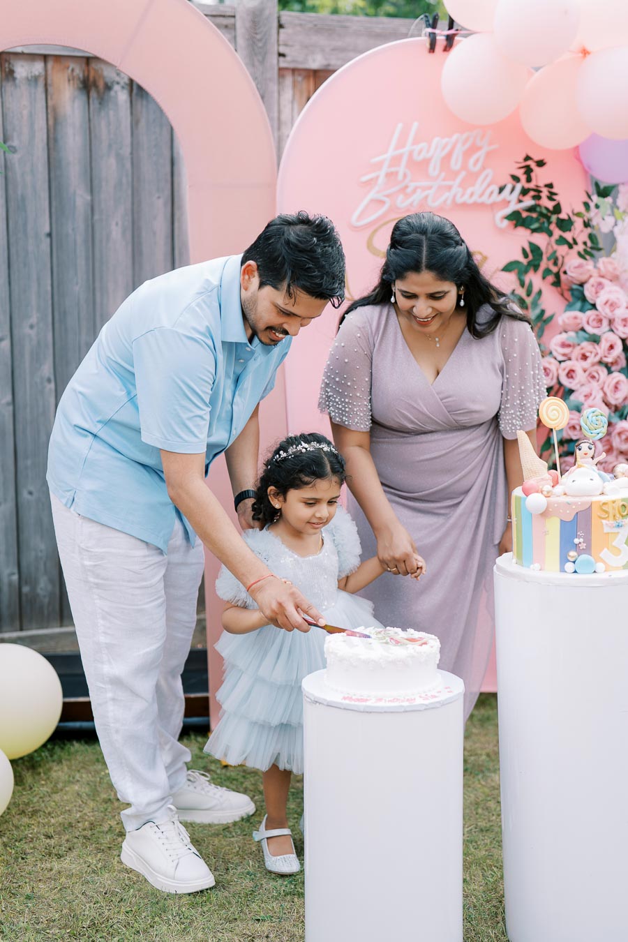 Family celebrating a child's birthday outdoors with themed decorations and a pastel cake, surrounded by pink and floral decor.