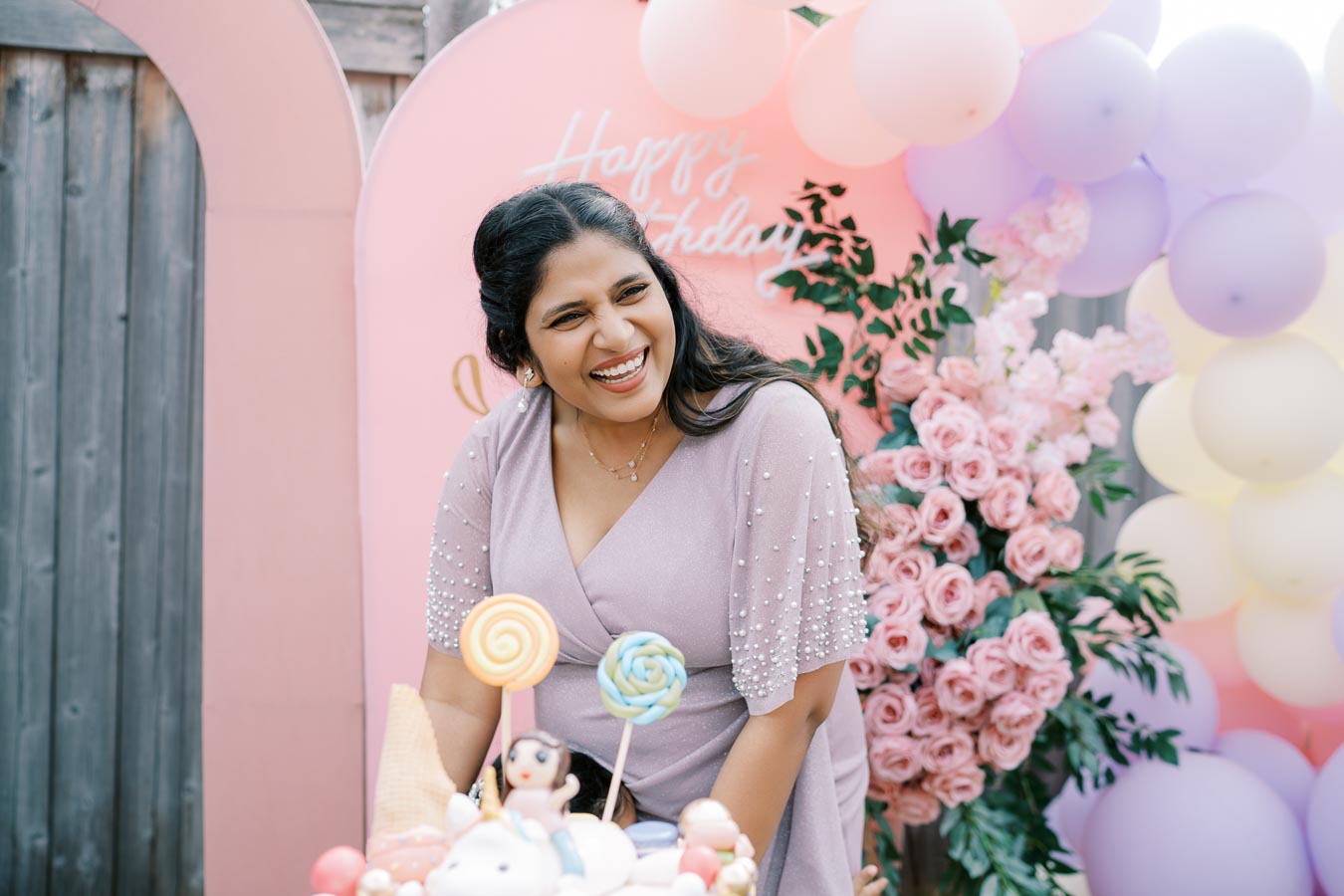 A woman in a lavender dress smiling at a birthday party with lollipop decorations, surrounded by pink roses and pastel balloons.