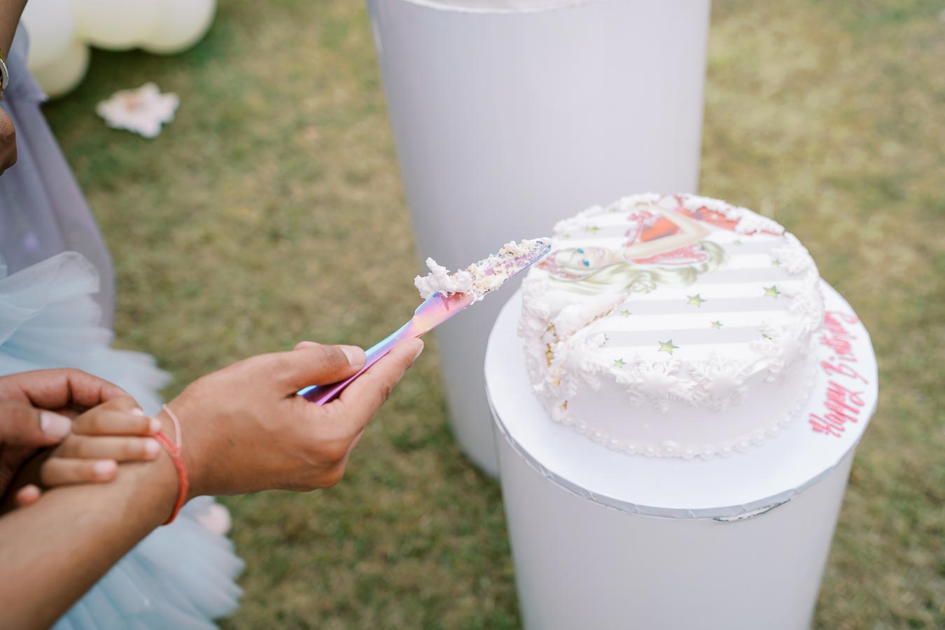 A person holding a colorful cake serving knife prepares to cut a white frosted birthday cake with decorative icing and a character design on top, set outdoors on a sunny day.