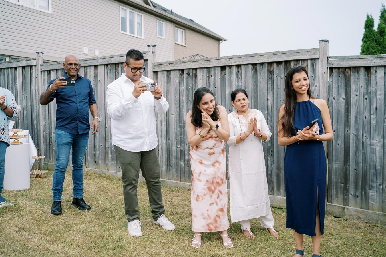 Group of people gathered in a backyard, smiling and taking photos with smartphones, during a casual outdoor gathering.