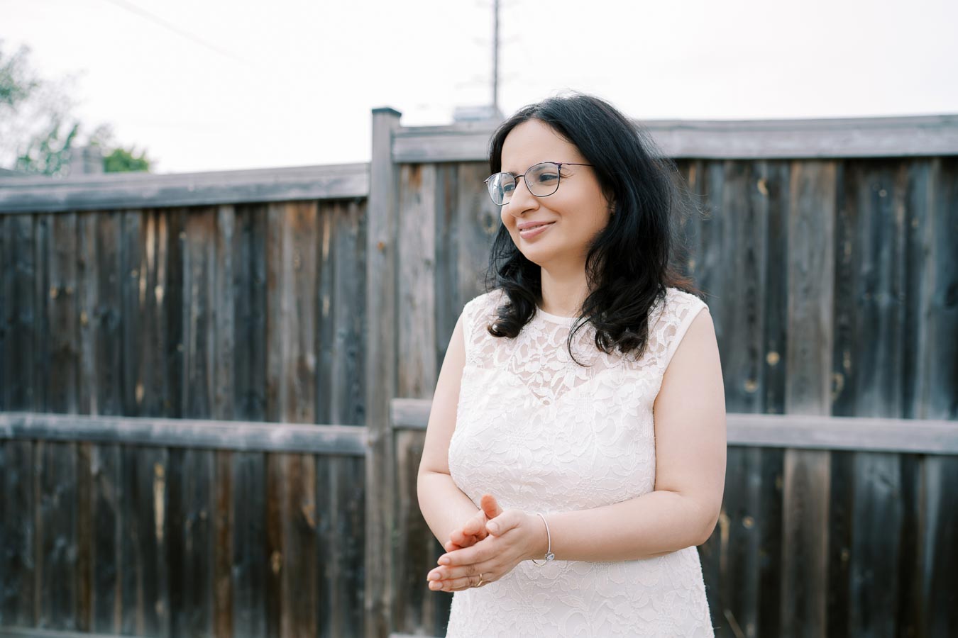 Woman in white lace dress smiling outdoors near a wooden fence.