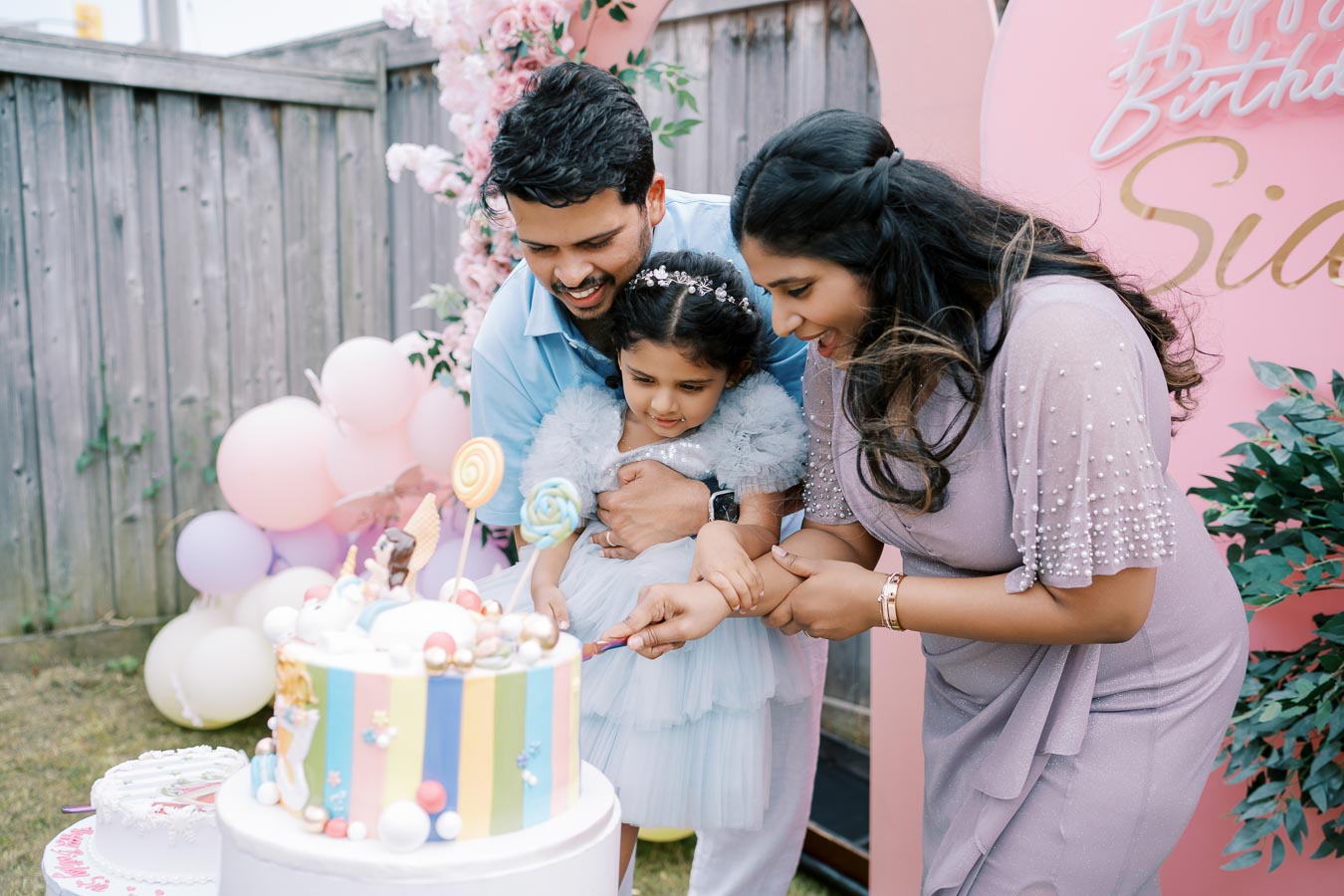 A joyful family celebrating a young girl's colorful birthday party, with parents helping her cut a large, vibrant cake adorned with lollipops, in a festive backyard setting decorated with balloons and flowers.