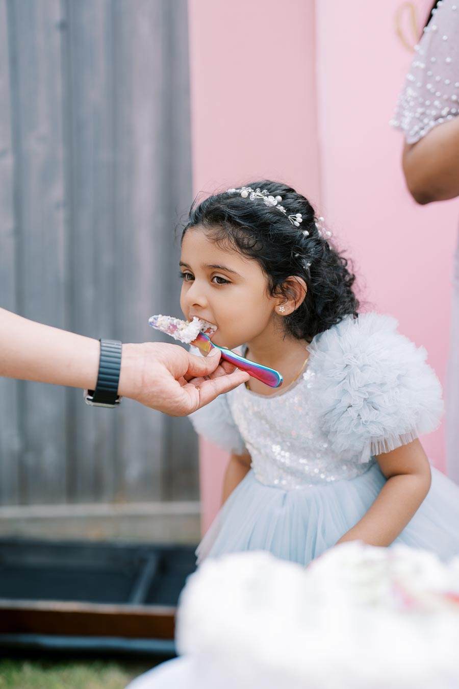 Little girl in a sparkling dress being fed cake with a colorful spoon at a party.