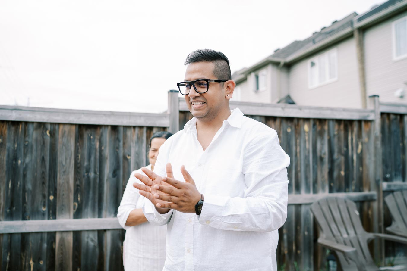 A man in a white shirt smiling and clapping in an outdoor setting with a wooden fence and patio chairs in the background.