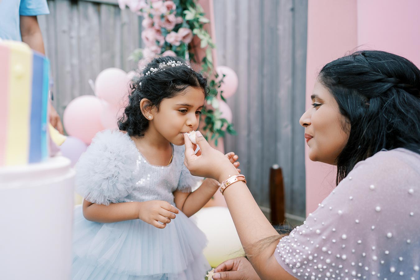 A mother feeds her young daughter a piece of cake at a colorful birthday party, surrounded by pink balloons and floral decorations.