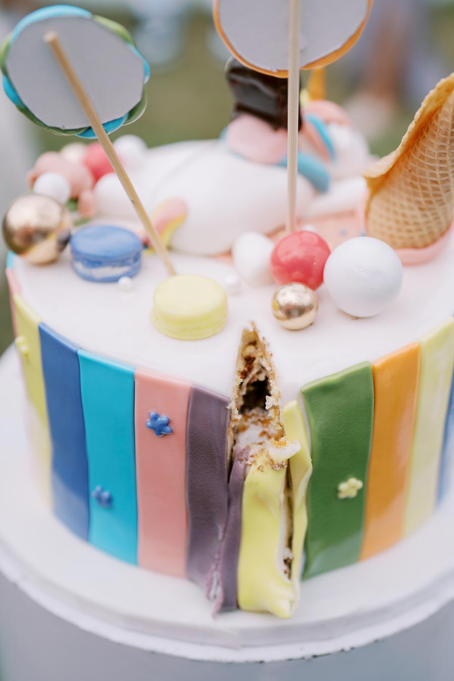 Colorful birthday cake with rainbow stripes, topped with macarons, gold spheres, and an ice cream cone, featuring a slice cut out. Perfect for a festive celebration or party theme.