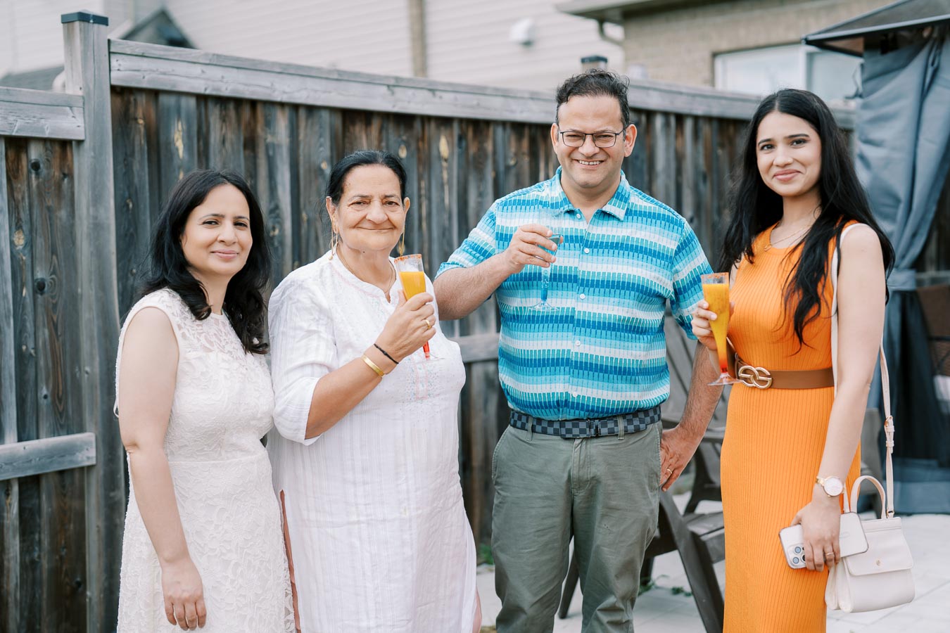 Four people smiling and holding drinks while posing for a photo outdoors, standing in front of a wooden fence.