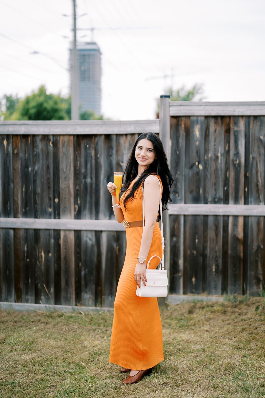 A woman in an orange dress stands outdoors, holding a drink and a white handbag, smiling in front of a wooden fence.