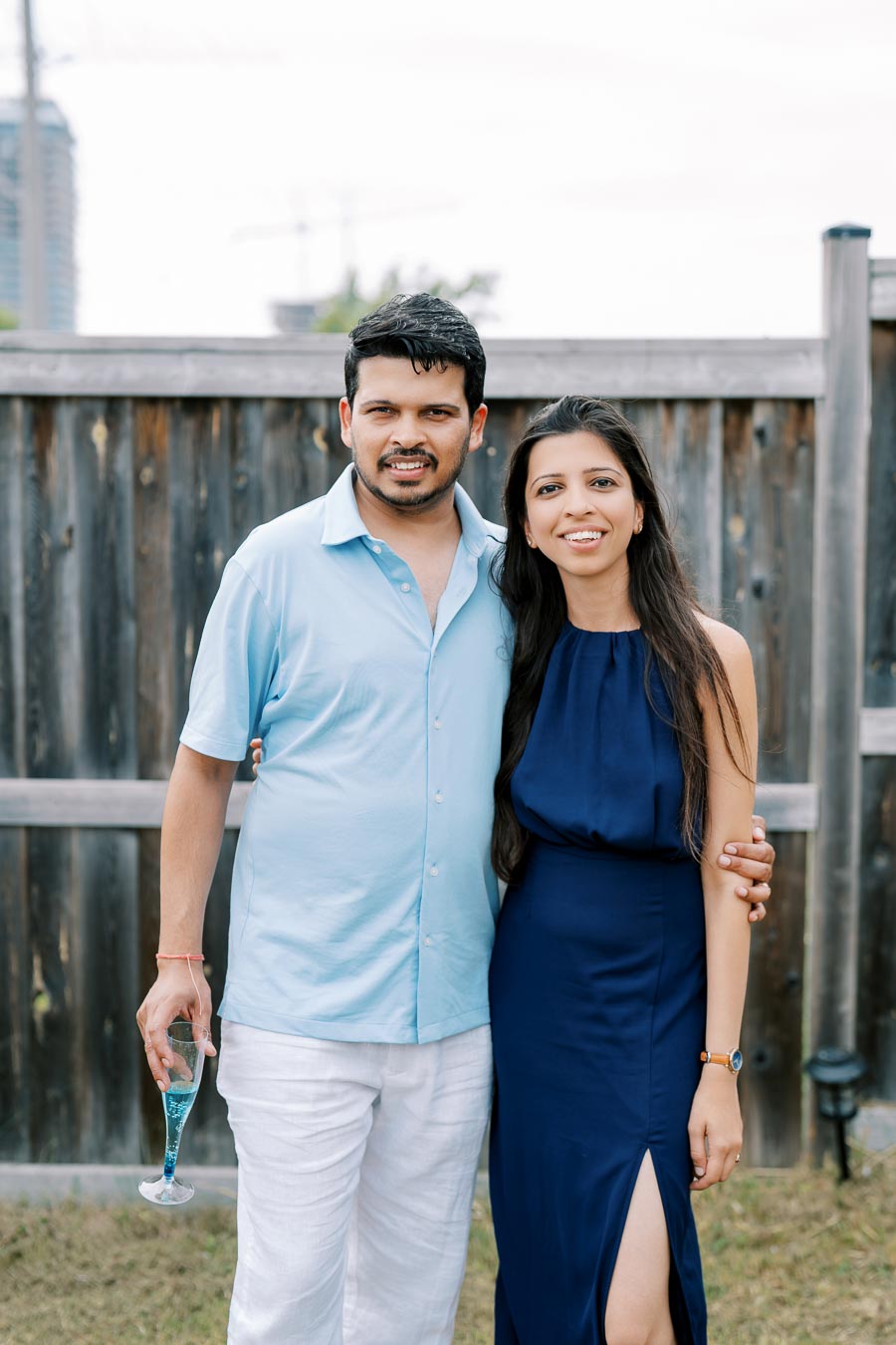 A couple wearing blue outfits posing together outside in front of a wooden fence, with the man holding a glass of blue liquid.