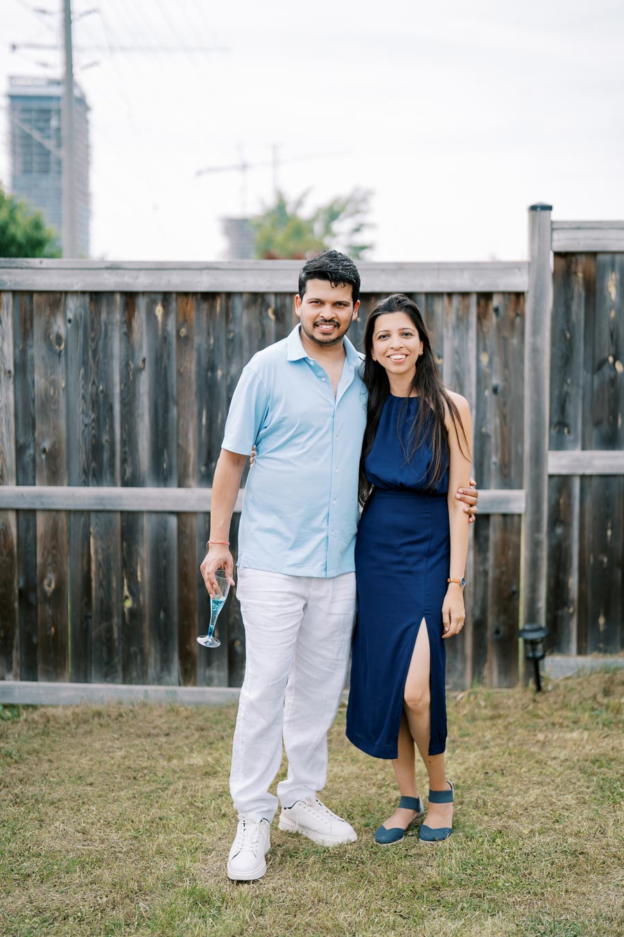 A smiling couple posing together outdoors in summer attire, standing in front of a wooden fence with a city skyline visible in the background. The man is holding a drink, and they appear relaxed and happy.