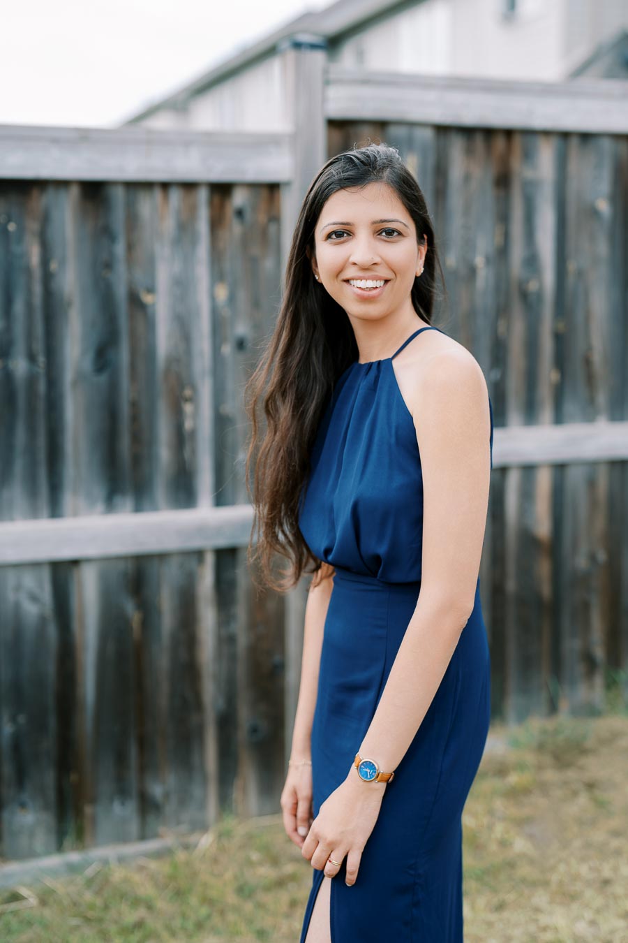 Smiling woman with long hair in a blue dress standing in front of a wooden fence outdoors.