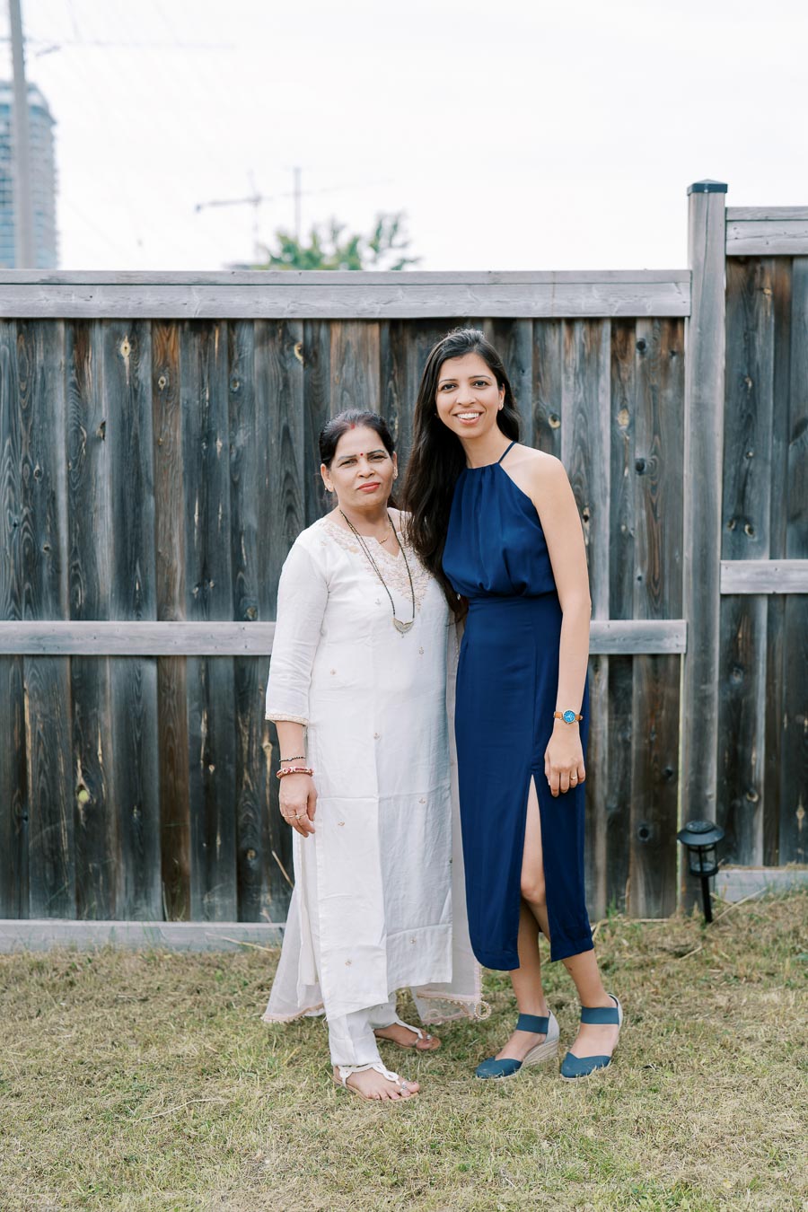 Two women smiling in front of a wooden fence, one wearing a blue dress and the other in a white traditional outfit, standing on grass.