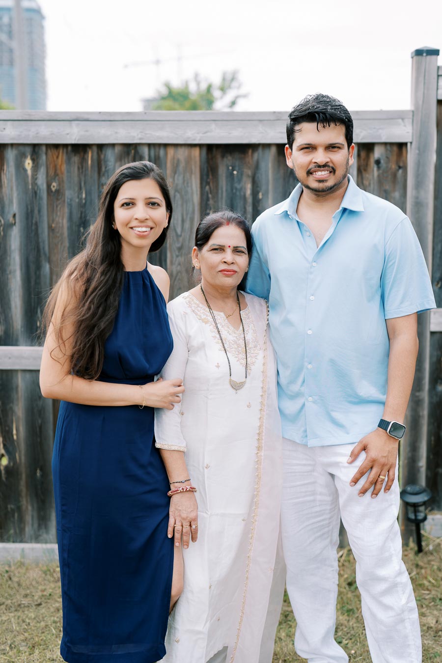 Family portrait in a backyard setting with a wooden fence, featuring a woman in a navy dress, an elder woman in a white traditional outfit, and a man in a light blue shirt and white pants, all smiling.