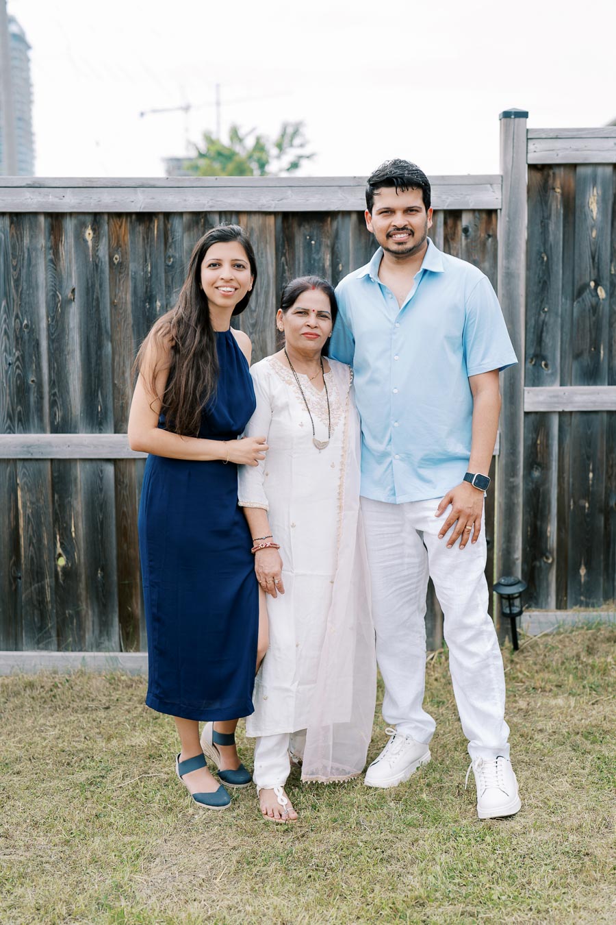 Family portrait outdoors with smiling individuals in front of a wooden fence, featuring a woman in a navy dress, a central figure in a traditional white outfit, and a man in a light blue shirt and white pants.