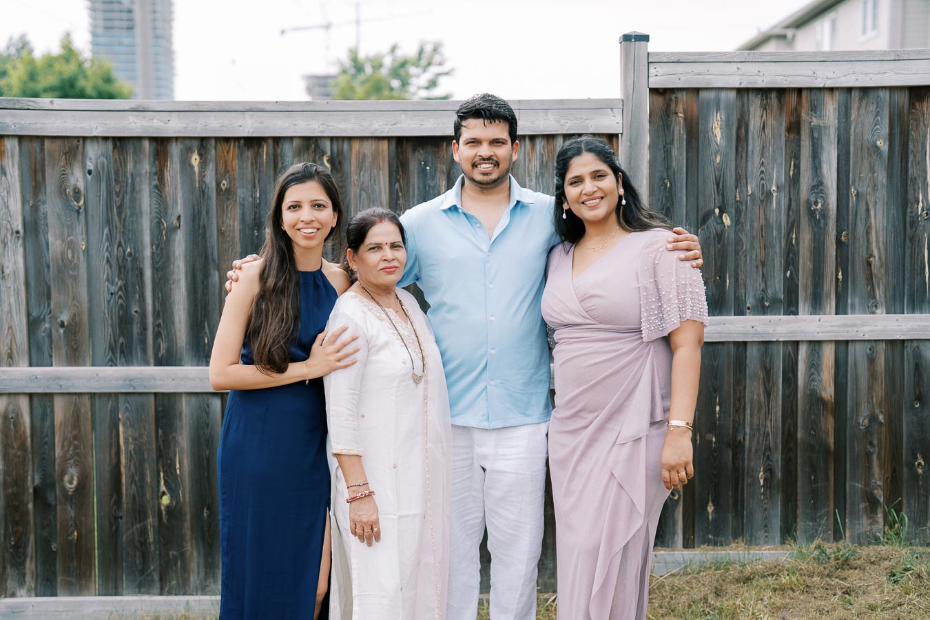 A family standing together in front of a wooden fence, featuring two women in dresses, a man in a light blue shirt, and an older woman in traditional attire, all smiling outdoors.