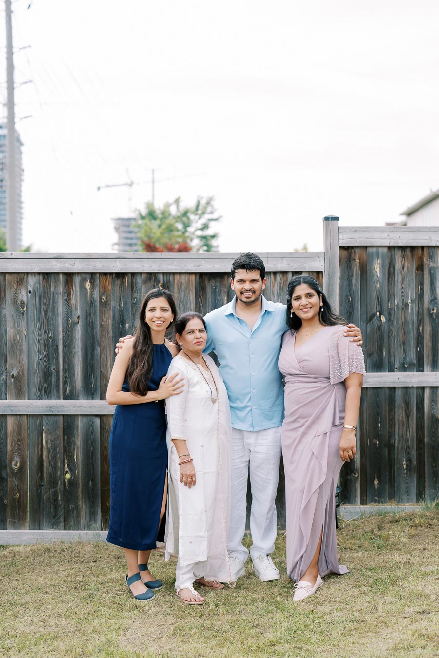 A family group photo in a backyard setting, featuring four people standing in front of a wooden fence. The individuals are dressed in summer attire, with a smiling woman in a navy dress, an older woman in a white outfit, a man in a light blue shirt, and a woman in a lavender dress. The background includes trees and a hint of construction